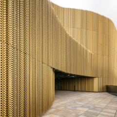 Curved building facade with perforated golden metal panels, creating a modern, textured surface. The ground is tiled, and the sky is partly cloudy in the background.