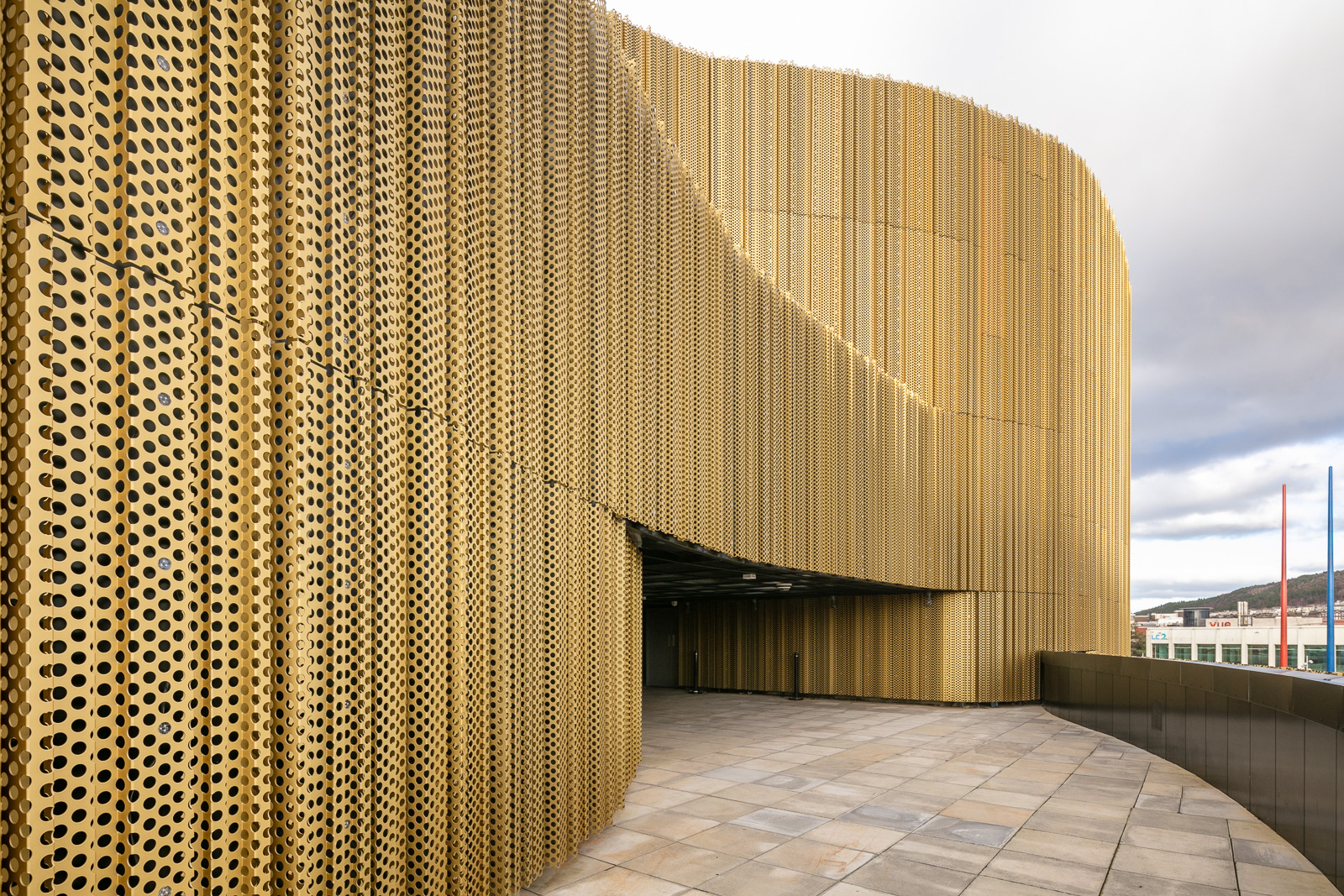Curved building facade with perforated golden metal panels, creating a modern, textured surface. The ground is tiled, and the sky is partly cloudy in the background.