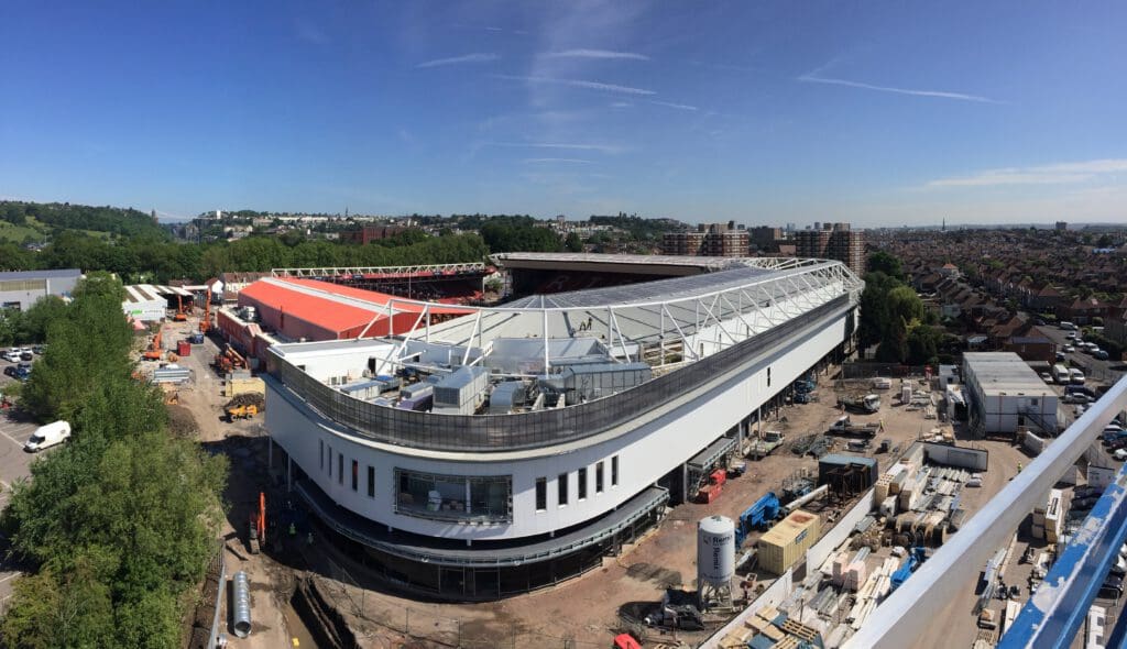 A panoramic view of a large stadium under construction, with cranes, construction materials, and machinery around. In the background are houses, trees, and a blue sky with wispy clouds.