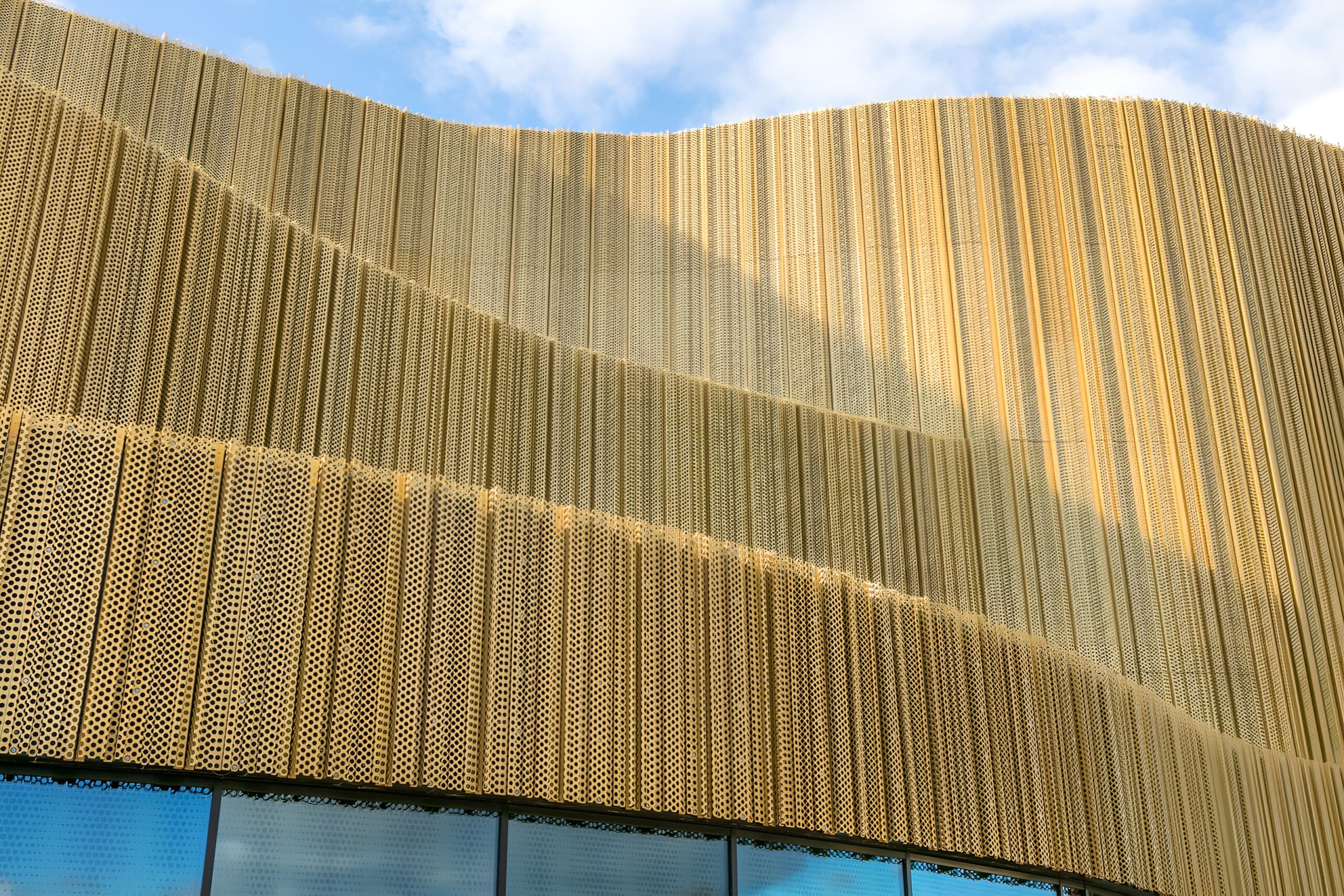 A modern building facade with wavy golden panels featuring a perforated pattern, reflecting sunlight, and a row of glass windows at the bottom. Blue sky with clouds is visible above.