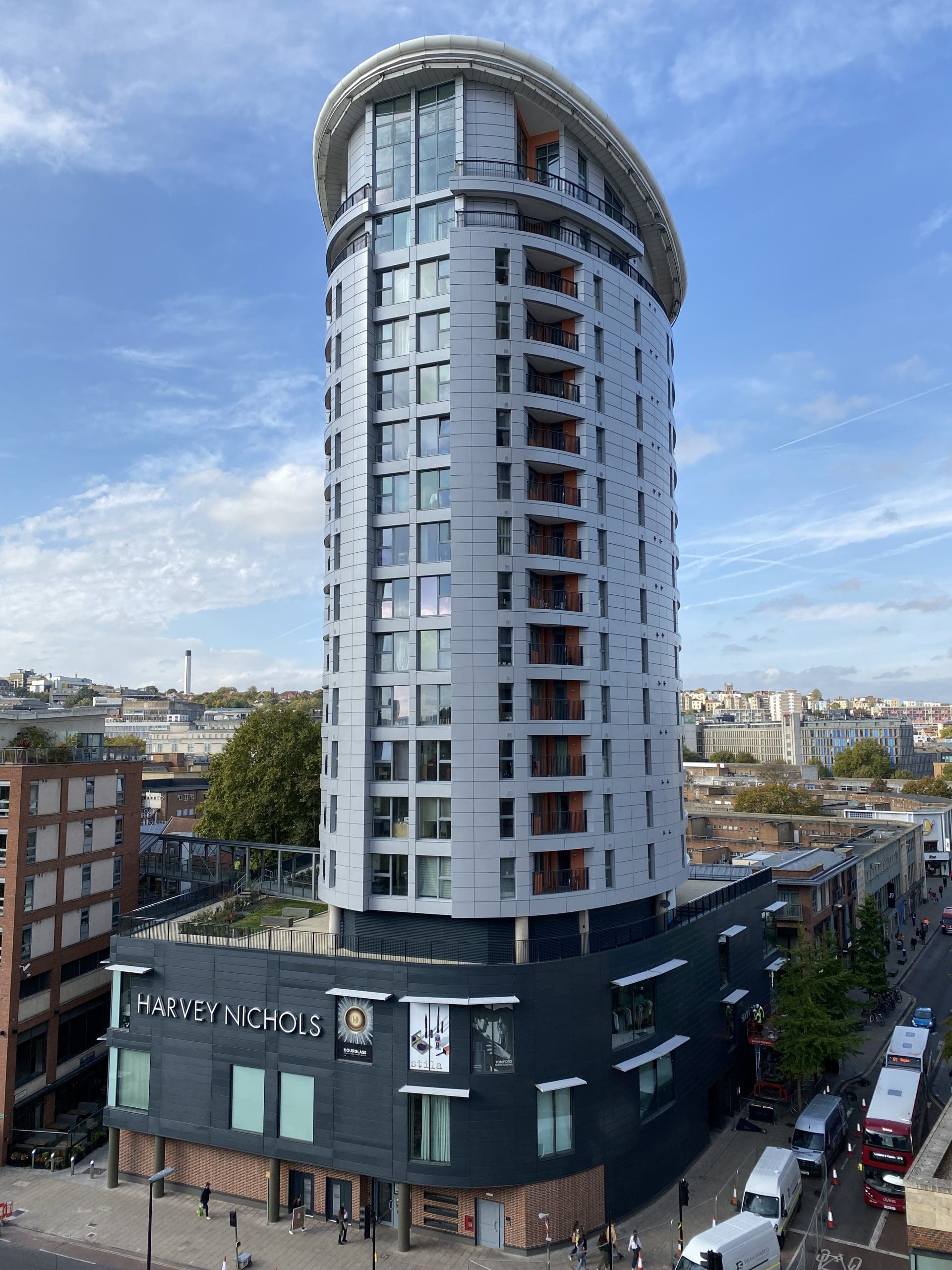 A tall, modern cylindrical building with many windows rises above a Harvey Nichols store. The sky is blue with scattered clouds, and people and vehicles move along the surrounding streets.