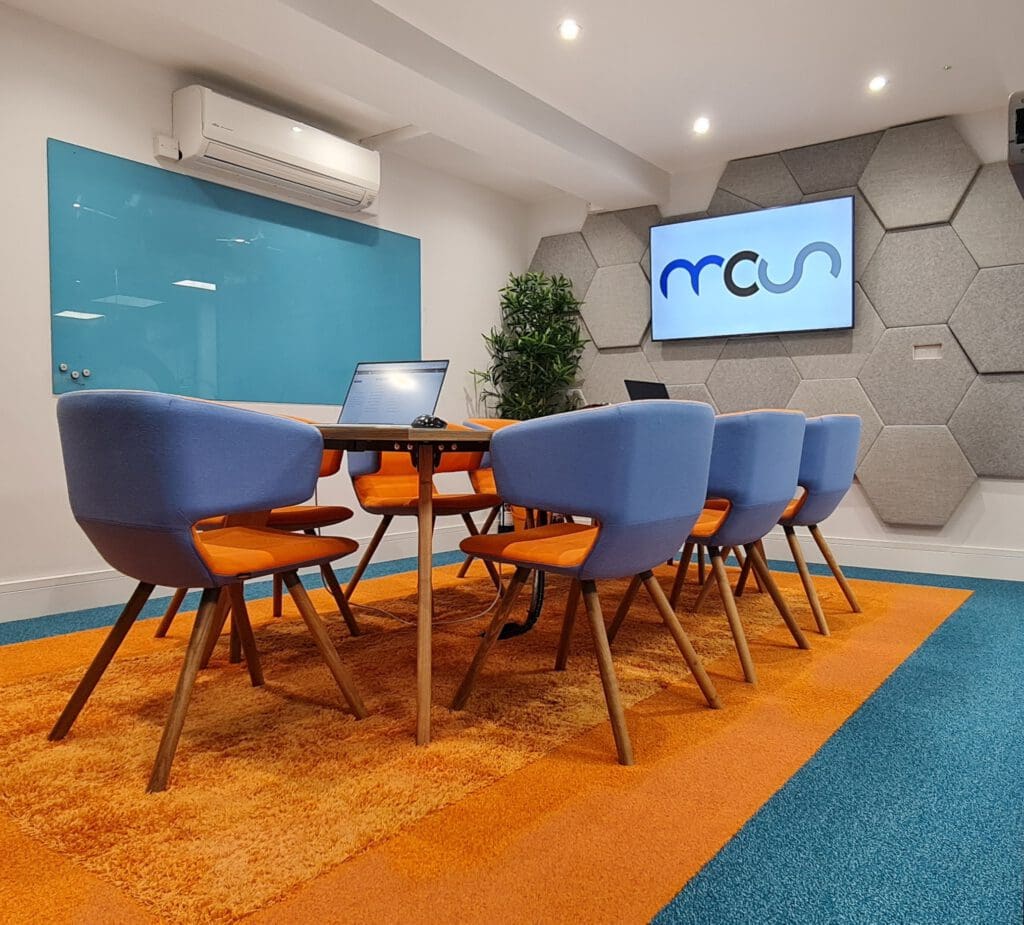 Modern conference room with a long wooden table, blue and orange chairs, a wall-mounted screen displaying "mrsa", a blue glass board, and geometric wall panels. Orange and blue carpet covers the floor.