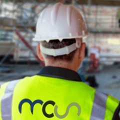 A construction worker wearing a white hard hat and a yellow reflective vest with a blue and green logo stands facing scaffolding at a building site. The background is blurred.