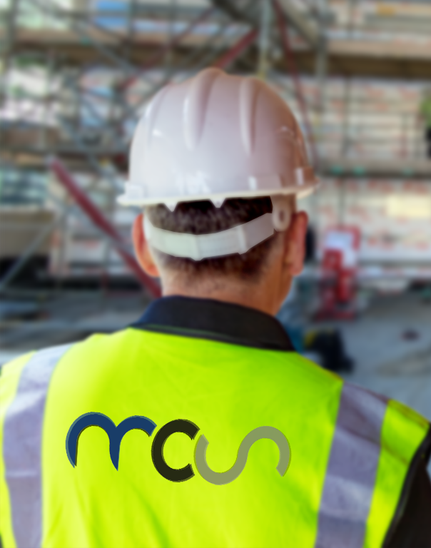A construction worker wearing a white hard hat and a yellow reflective vest with a blue and green logo stands facing scaffolding at a building site. The background is blurred.
