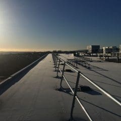 A flat rooftop with metal railings and large HVAC units, lit by the low morning or evening sun, overlooking a distant landscape under a clear blue sky.