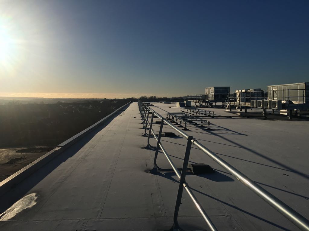 A flat rooftop with metal railings and large HVAC units, lit by the low morning or evening sun, overlooking a distant landscape under a clear blue sky.