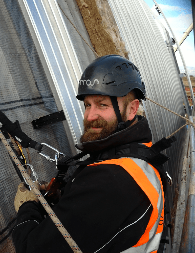 A bearded construction worker wearing a black helmet and orange safety vest is secured with climbing gear while working on the side of a metal-clad building, looking back and smiling at the camera.