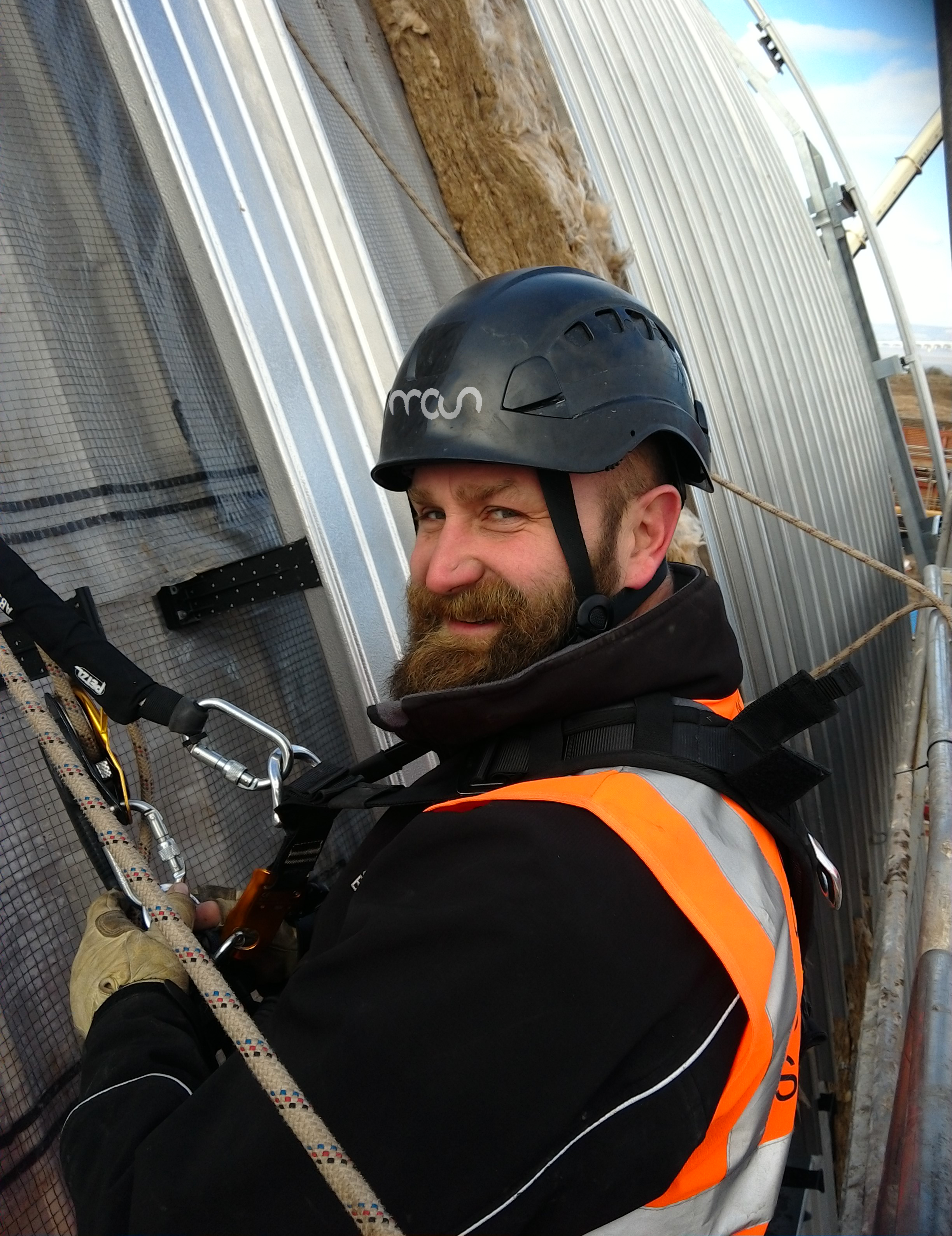 A bearded construction worker wearing a black helmet and orange safety vest is secured with climbing gear while working on the side of a metal-clad building, looking back and smiling at the camera.