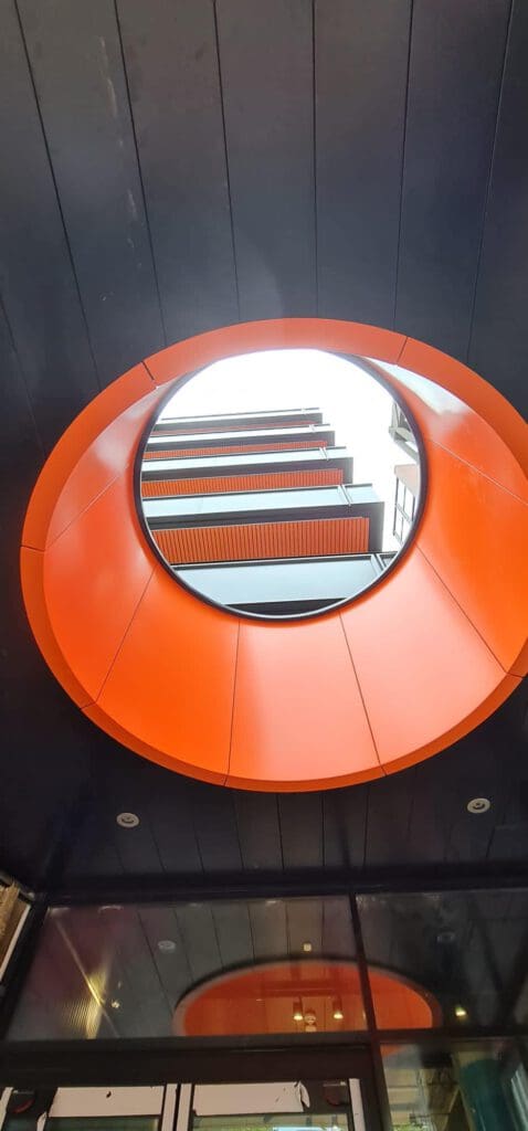 A view looking up through a large circular orange structure in the ceiling, revealing the exterior of a modern building with horizontal lines and glass balconies above.