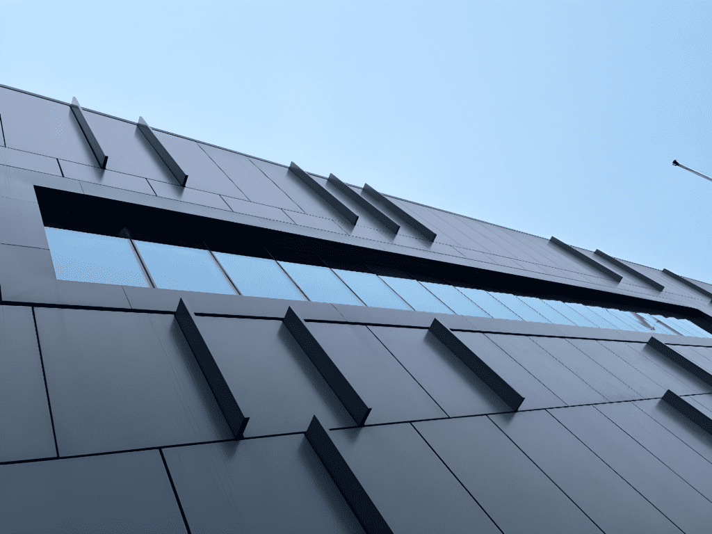 A modern building with sleek, gray, rectangular panels and angled architectural features, viewed from below against a clear blue sky. The design is minimalist and geometric, with reflective windows along one side.