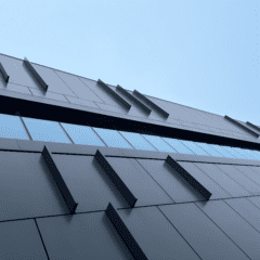 A modern building with sleek, gray, rectangular panels and angled architectural features, viewed from below against a clear blue sky. The design is minimalist and geometric, with reflective windows along one side.