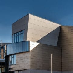 A modern building with geometric, angular design, featuring light-colored panels, large windows, and sharp shadows cast by the sun under a clear blue sky.