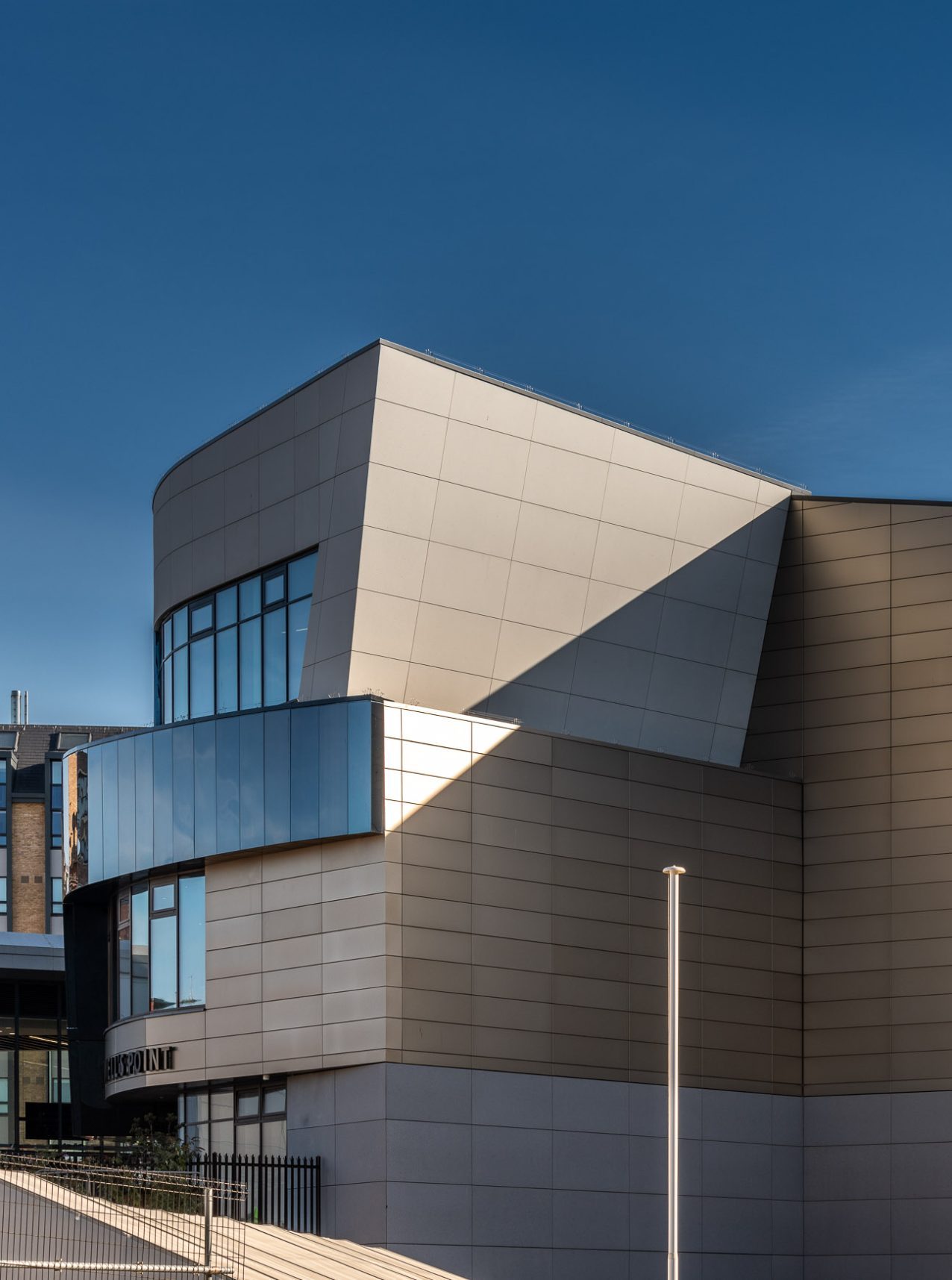 A modern building with geometric, angular design, featuring light-colored panels, large windows, and sharp shadows cast by the sun under a clear blue sky.
