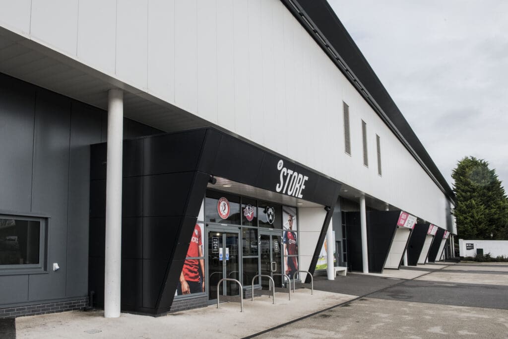 The image shows the exterior of a modern sports stadium store with glass doors, team logos, and merchandise posters on display, set under a black and white overhang. Bike racks are positioned in front of the entrance.