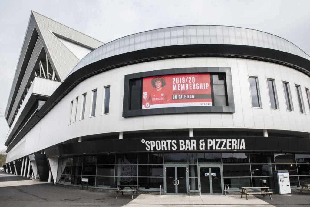 Modern building with large windows and a curved facade, featuring a sign for "SPORTS BAR & PIZZERIA" and an electronic billboard above advertising 2019/20 memberships.