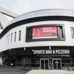 Modern building with large windows and a curved facade, featuring a sign for "SPORTS BAR & PIZZERIA" and an electronic billboard above advertising 2019/20 memberships.