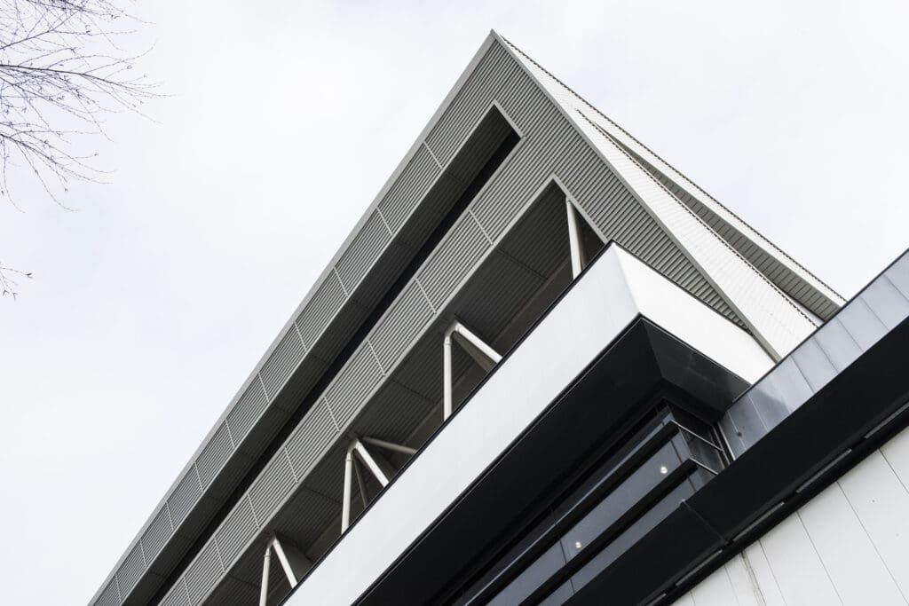 A modern building with angular, geometric lines and a mix of gray, white, and black panels, viewed from a low angle against a cloudy sky.