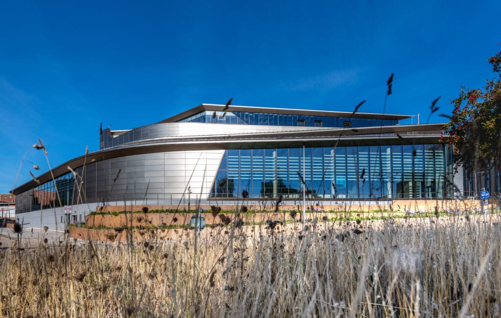 A modern building with a curved metal and glass facade, viewed from behind tall dry grass under a clear blue sky.