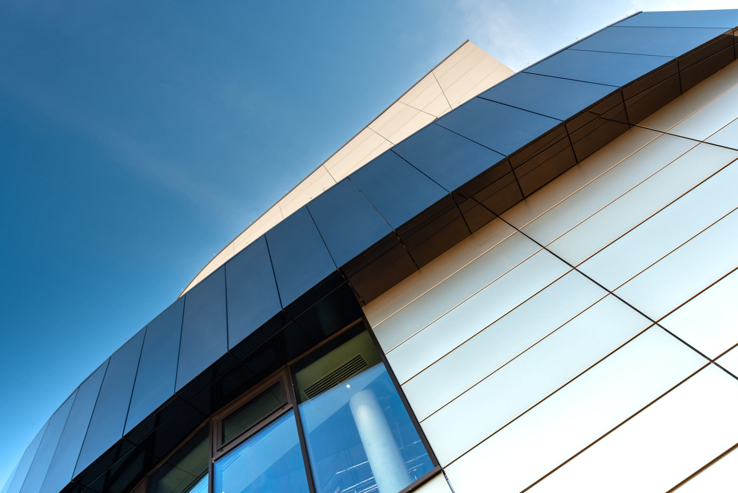 Close-up view of a modern building with reflective glass and metal panels, captured from a low angle against a clear blue sky. Geometric lines and sharp angles dominate the architecture.