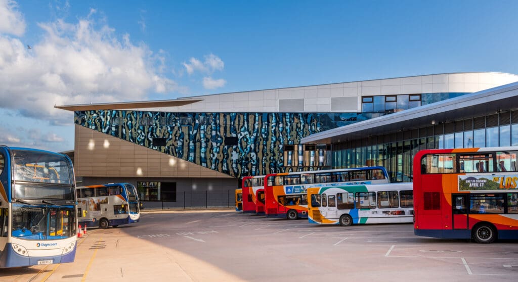Buses parked at a modern bus station with a large building featuring blue and white abstract glass artwork, under a blue sky with scattered clouds.