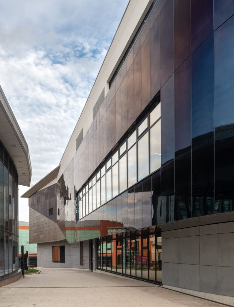 Modern glass and steel building with large reflective windows under a partly cloudy sky, showing clean lines and geometric shapes; paved walkway in the foreground.