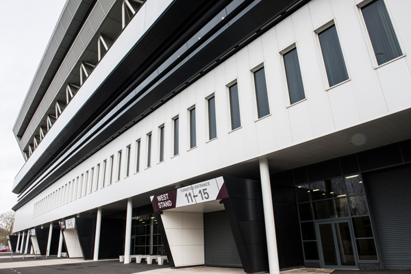Exterior view of a modern stadium building with large windows, bold geometric lines, and overhanging upper levels. Signs indicate the "West Stand" and entrances/conferences 11-15.