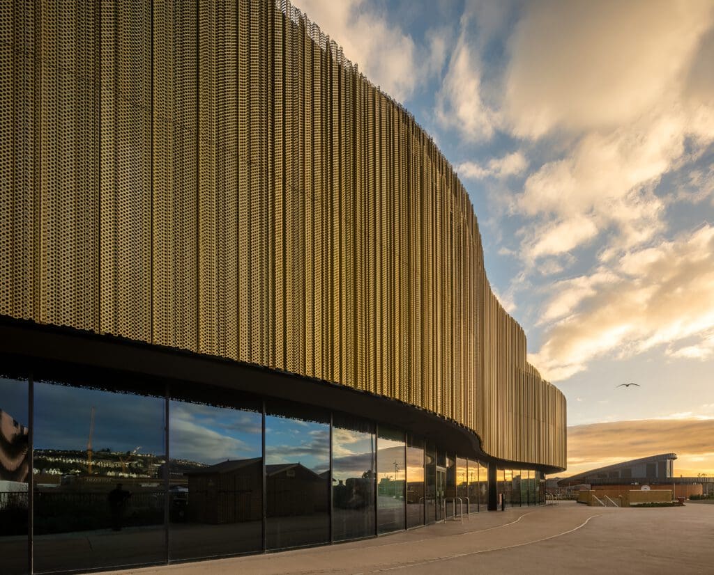 A modern building with a curved gold metal facade and large glass windows, reflecting the cloudy sky during sunset, creating a warm and dramatic atmosphere.