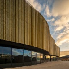 A modern building with a curved gold metal facade and large glass windows, reflecting the cloudy sky during sunset, creating a warm and dramatic atmosphere.