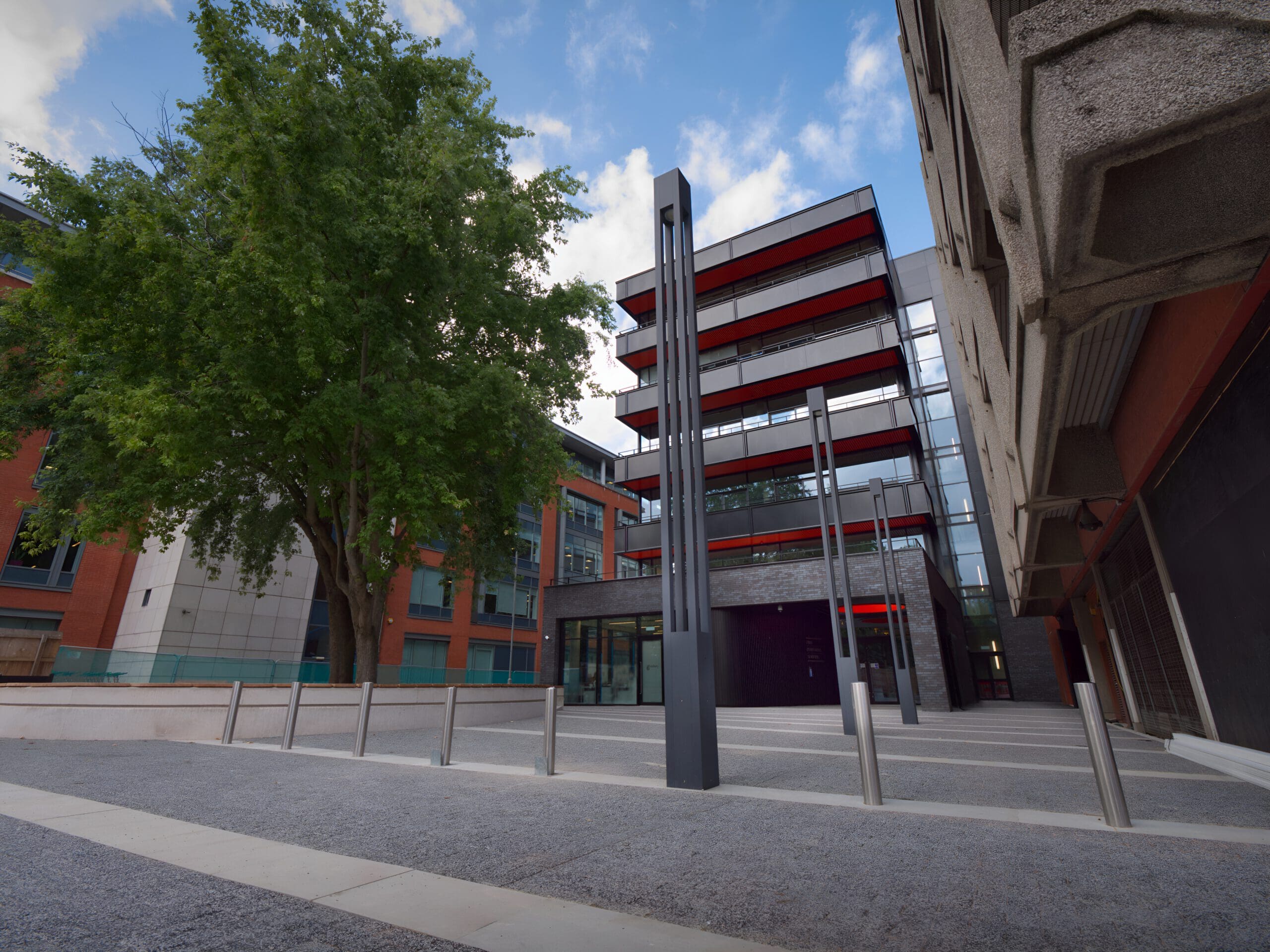 A modern building with red and black accents stands behind a tree, metal poles, and a gravel courtyard, with another concrete building on the right and a partly cloudy sky above.