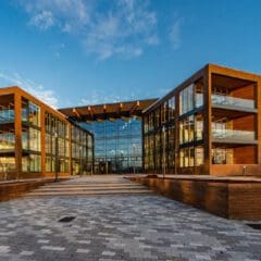 A modern, three-story glass and wood building with large balconies and a wide paved walkway leading to the entrance, photographed under a clear blue sky.