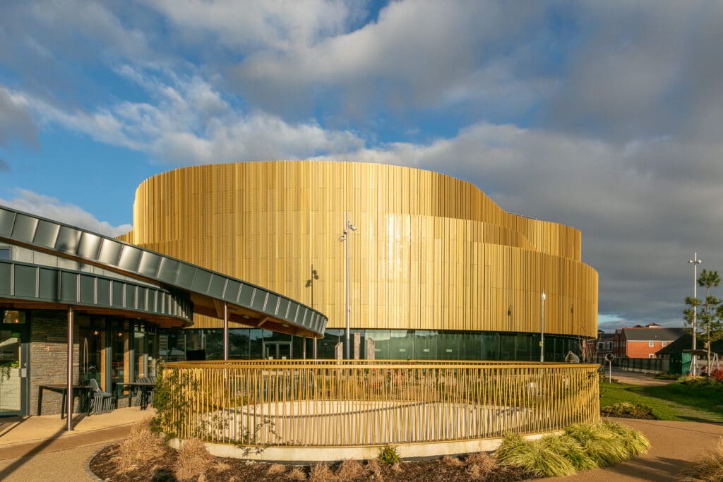 A modern building with curved golden metal exterior panels and large glass windows, set against a partly cloudy sky, with landscaped plants and a railing in the foreground.