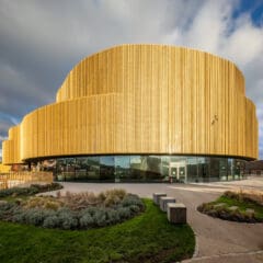 A modern, circular building with a golden, ribbed exterior and large glass windows at ground level, surrounded by landscaped greenery and a curved pathway under a partly cloudy sky.