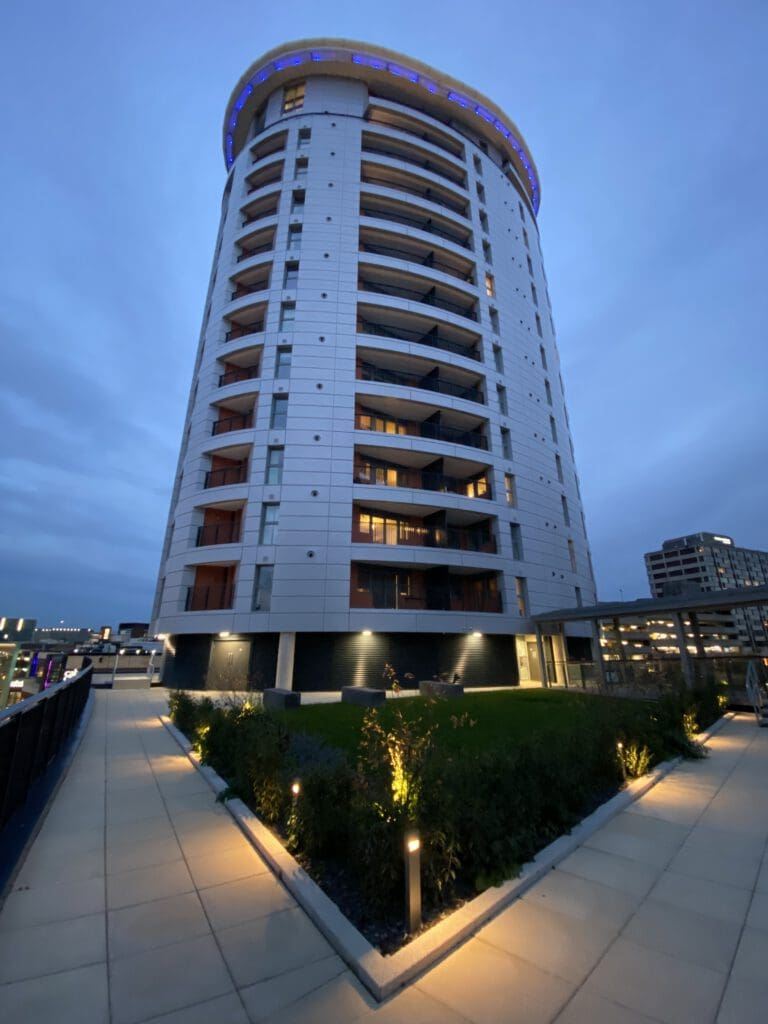 A modern, tall, circular apartment building with balconies, illuminated by blue lights at the roof, stands at dusk. The foreground shows a landscaped garden and a paved walkway lined with small lights.