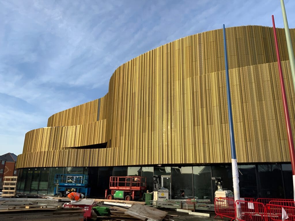 A modern building with a curved, golden metal facade under construction. Construction equipment, materials, and barriers are visible in front of large glass windows on the ground floor, with a blue sky overhead.