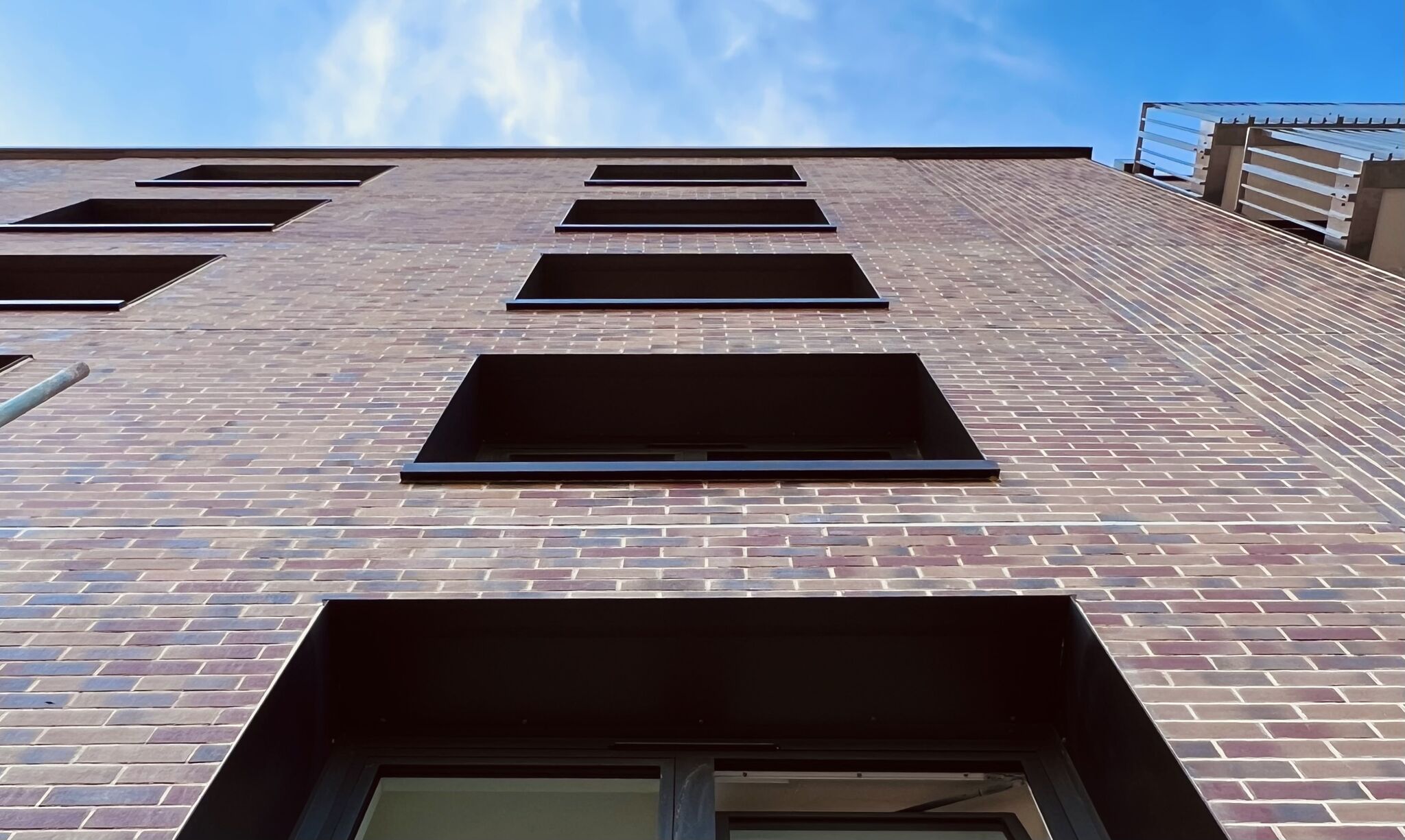 Looking up at a modern brick building with multiple rectangular windows, set against a partly cloudy blue sky. The perspective emphasizes the building's height and geometric lines.