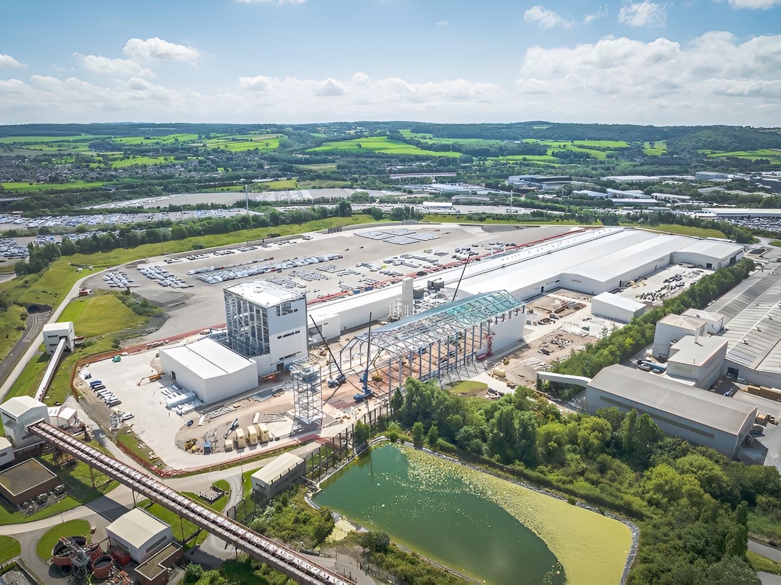 Aerial view of a large industrial complex under construction, with cranes, steel framework, white-roofed buildings, parking lots, green fields, trees, and a pond in the foreground under a partly cloudy sky.