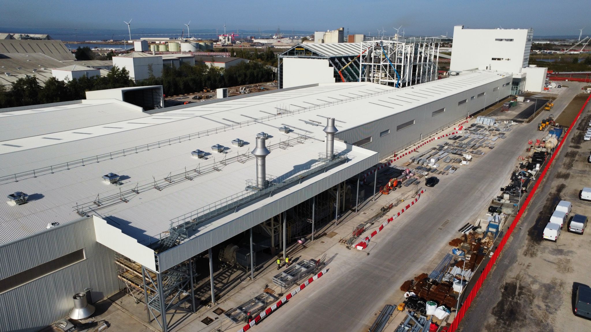 A large industrial facility under construction, with steel framework, white-roofed buildings, construction materials, trucks, and equipment visible; wind turbines and water are in the distant background.