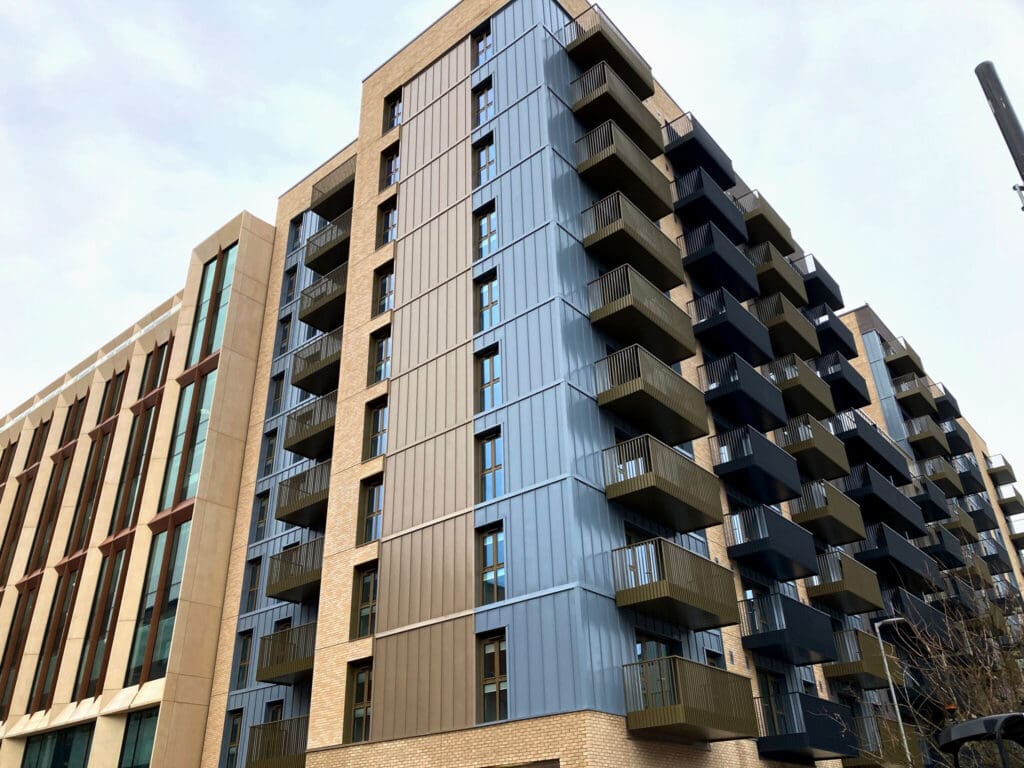 A modern apartment building with light brown brick and vertical panels in shades of blue and bronze, featuring multiple dark balconies and large windows, next to another building with a beige facade and tall windows.