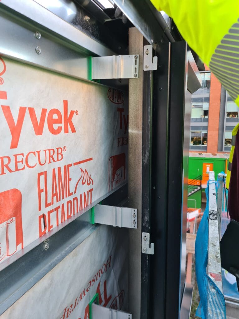 Close-up of a building under construction, showing Tyvek fire-retardant sheathing, metal brackets, and insulation layers. A worker in a yellow vest is visible on the right, with scaffolding and buildings in the background.