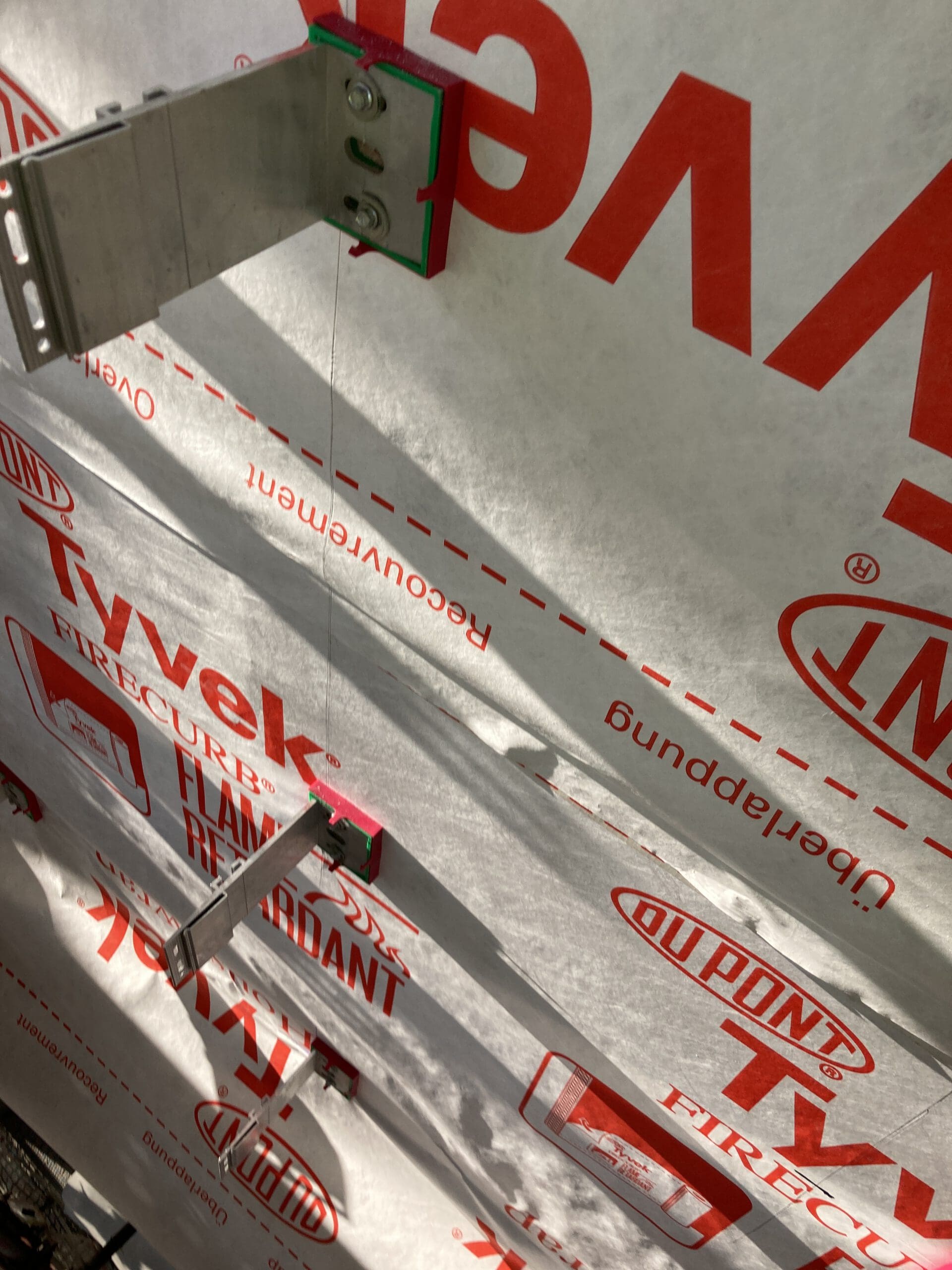 Metal brackets attached to a wall covered in white DuPont Tyvek building wrap, with red text and logos, under sunlight. The brackets cast shadows on the surface.