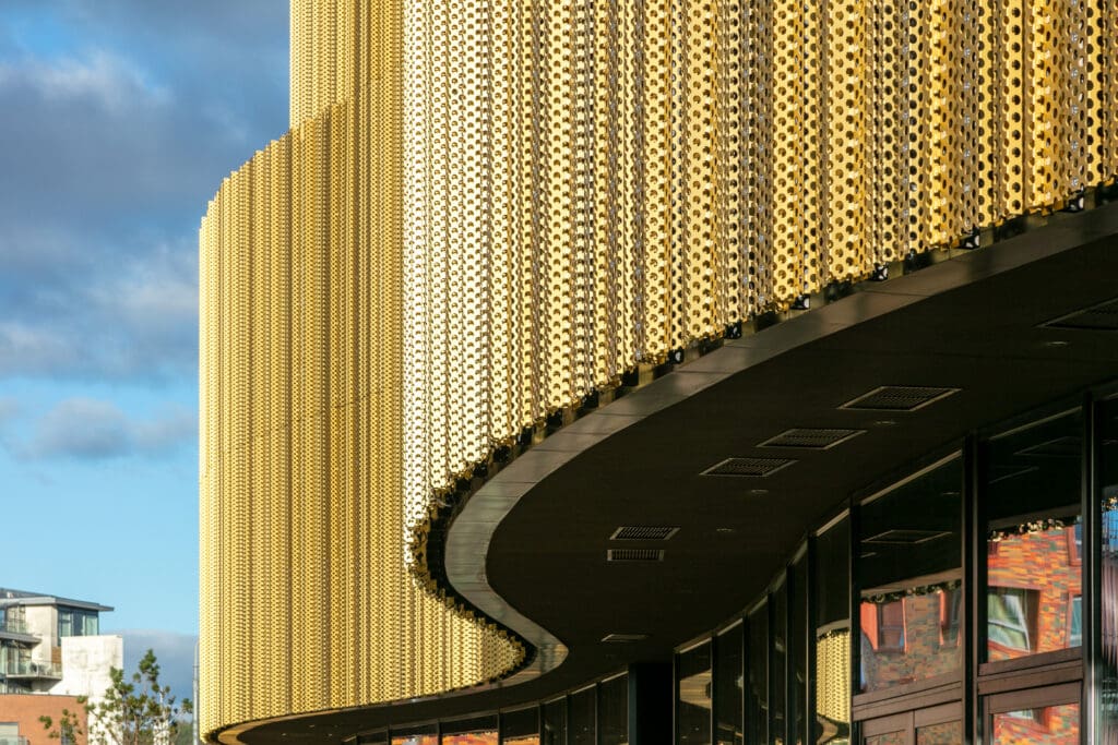 Close-up of a modern building with a curved facade covered in gold-colored, perforated metal panels, reflecting sunlight. The lower part features dark glass windows and overhangs, with part of the sky and nearby buildings visible.