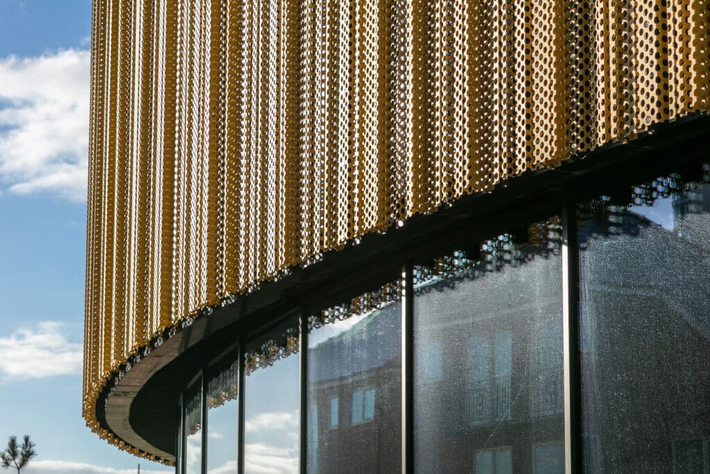 A close-up of a modern building with large glass windows and a golden perforated metal facade, reflecting sunlight against a blue sky with scattered clouds.