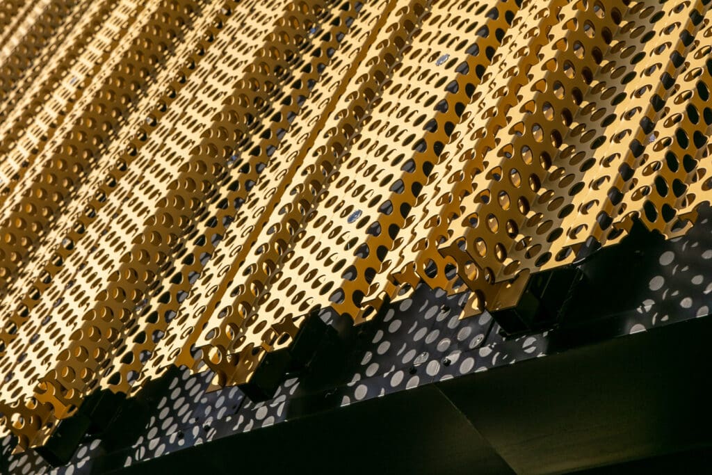 Close-up of a modern architectural facade featuring angled, gold perforated metal panels with circular holes, casting shadows on a black surface with white dots below.