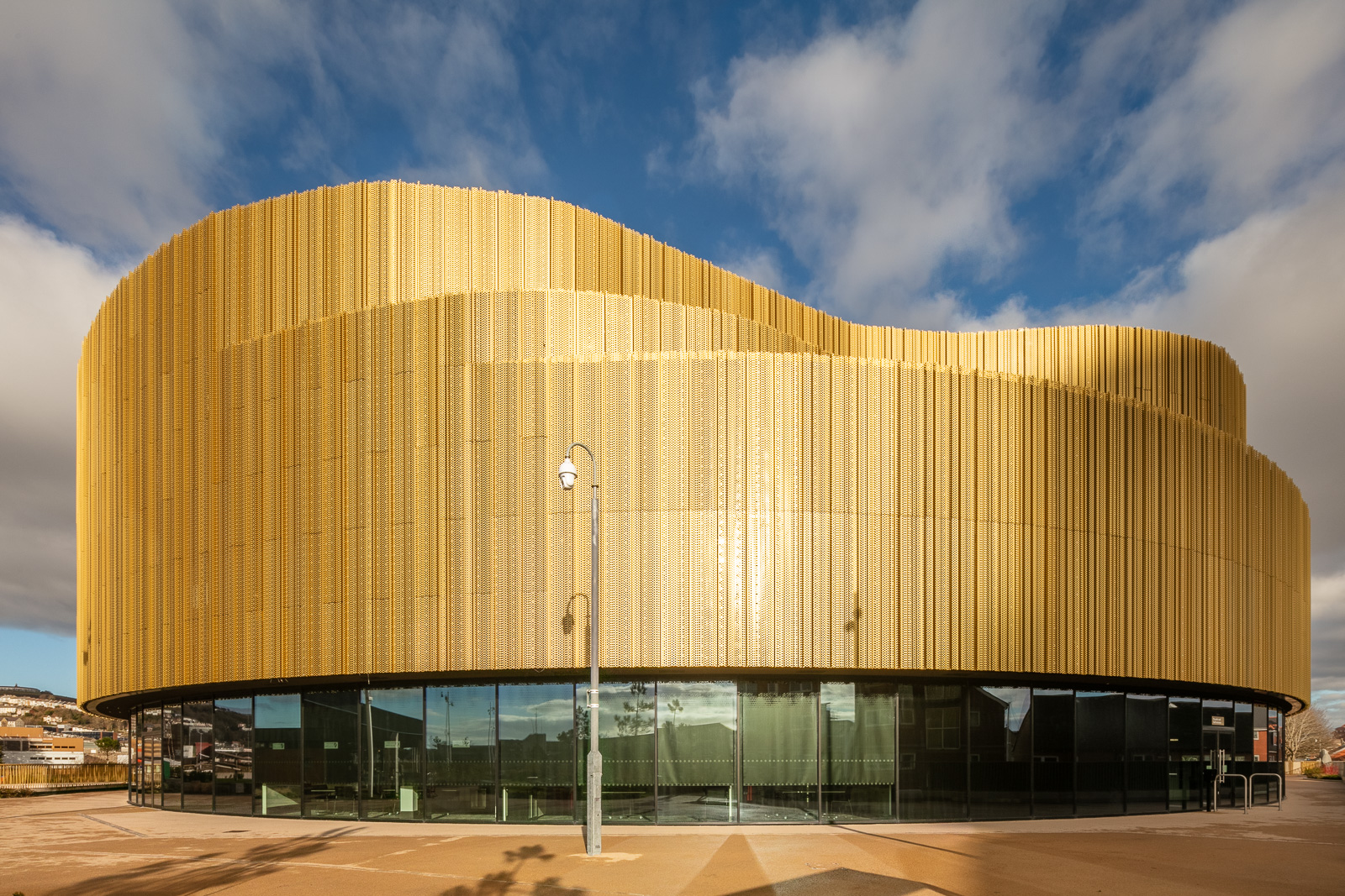 A modern building with a curving gold-colored exterior and large glass windows at the ground level, set against a blue sky with scattered clouds.