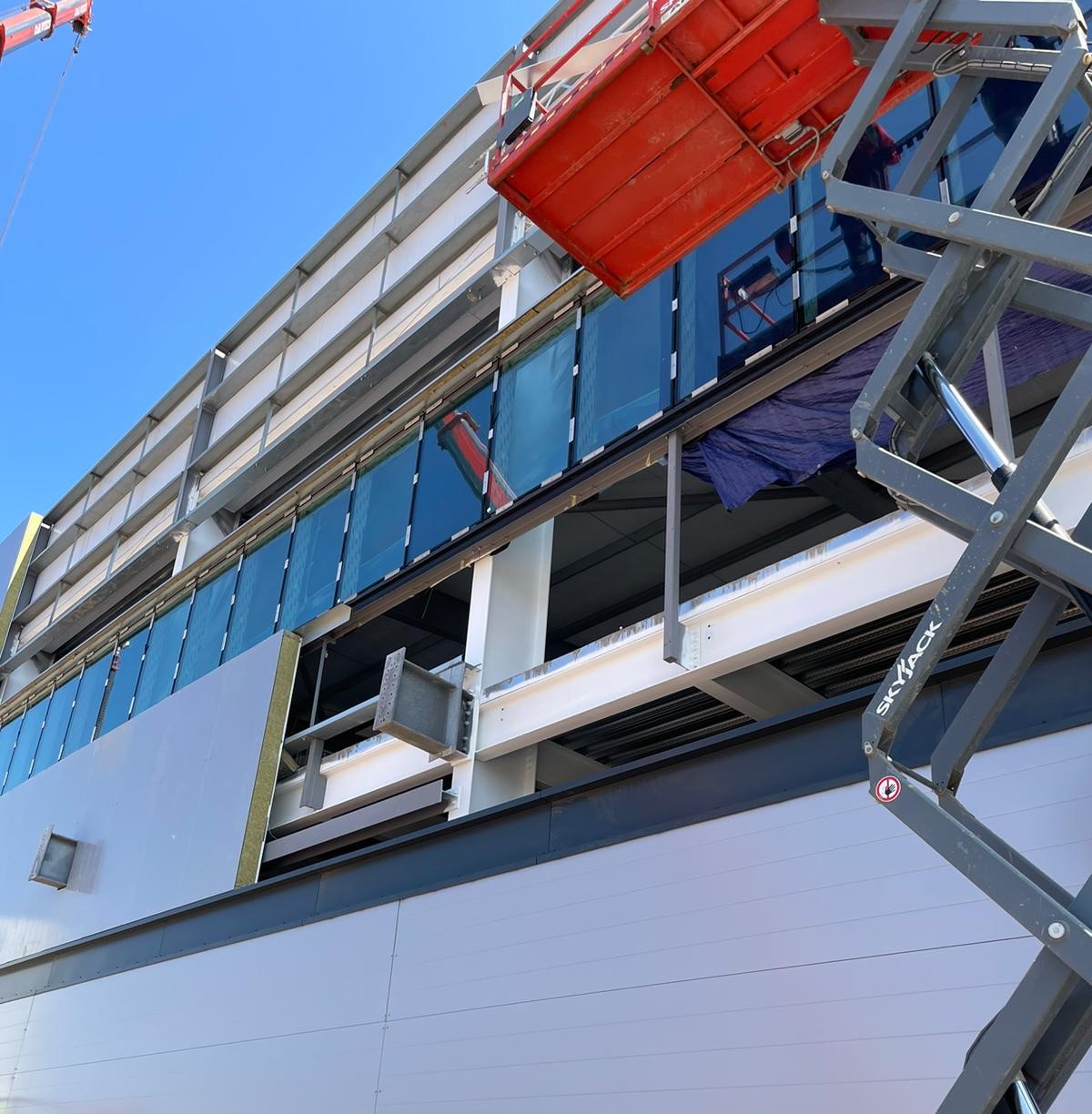 Construction workers on cherry pickers install glass panels on the exterior of a modern building under a clear blue sky. Some areas are unfinished, exposing metal framework and building materials.