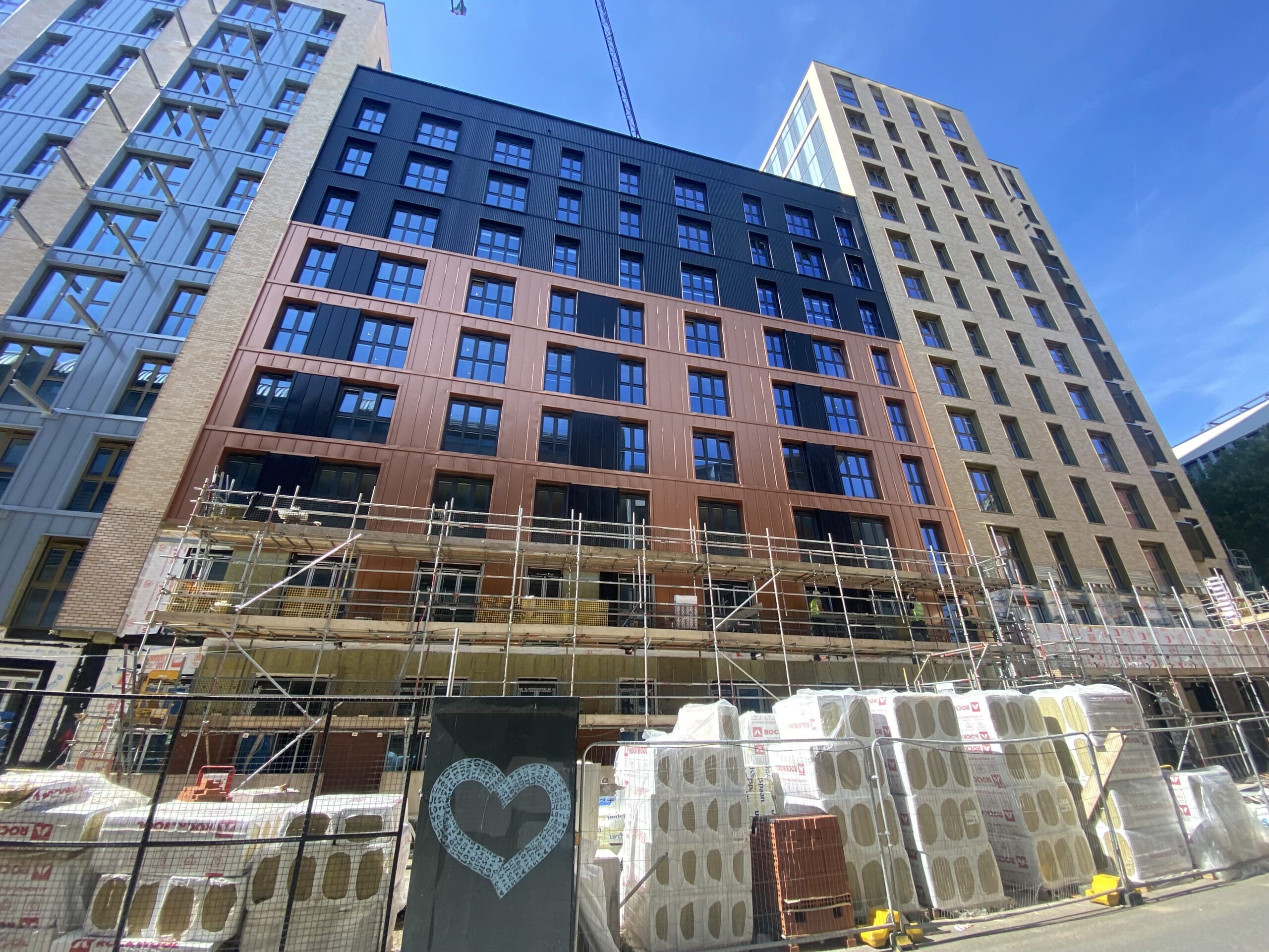 A multi-story building under construction with scaffolding and building materials stacked in front. A black panel with a white heart is visible in the foreground. The sky is clear and blue.