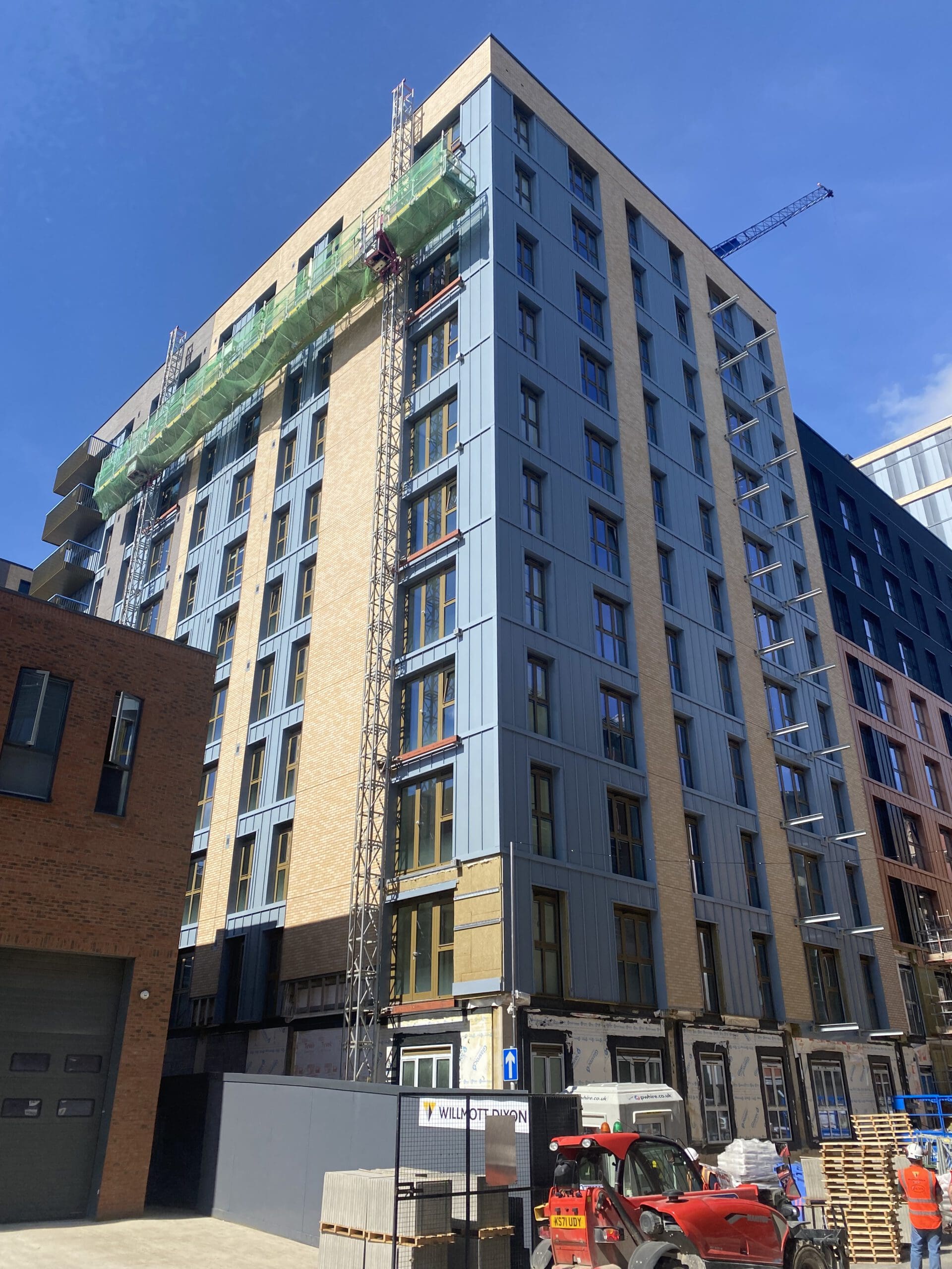 A mid-rise modern apartment building under construction, with scaffolding and green safety netting on the upper floors, construction materials and vehicles in front, and a clear blue sky above.