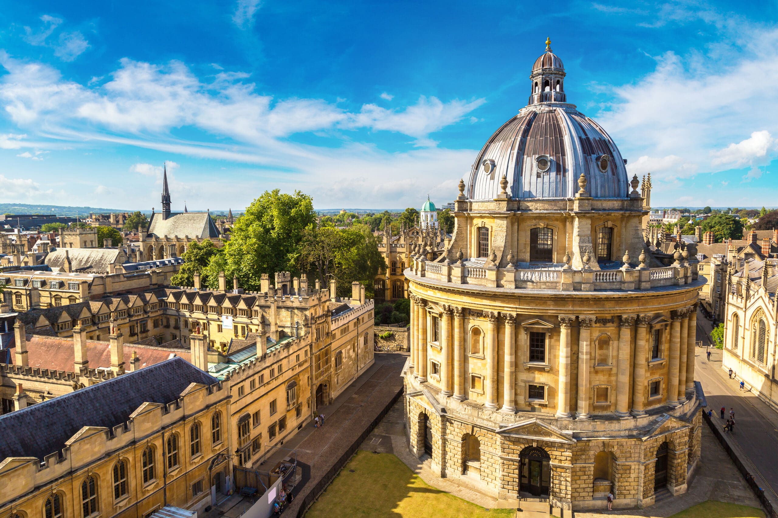 Aerial view of the Radcliffe Camera, a historic domed library building in Oxford, England, surrounded by old university buildings under a bright blue sky.