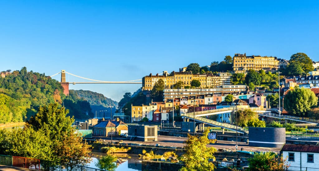 A wide view of Bristol, England, featuring colorful houses, lush green trees, and the Clifton Suspension Bridge spanning a deep gorge under a clear blue sky.