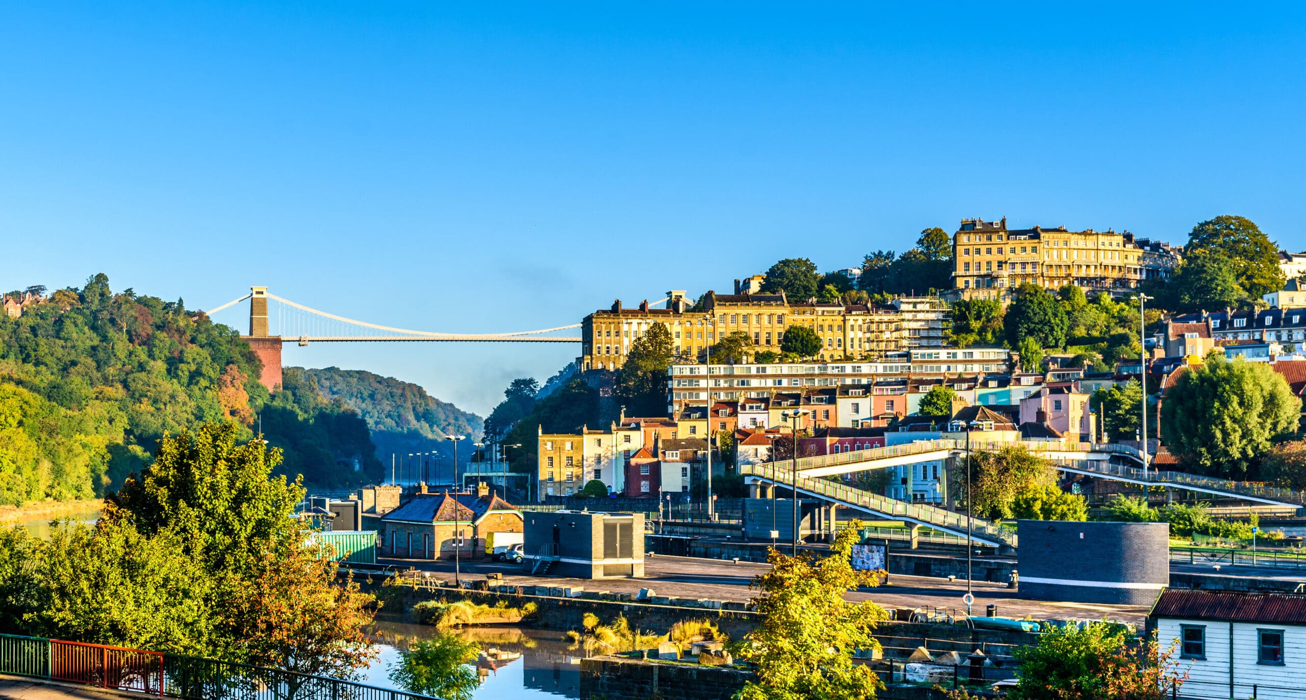 A wide view of Bristol, England, featuring colorful houses, lush green trees, and the Clifton Suspension Bridge spanning a deep gorge under a clear blue sky.